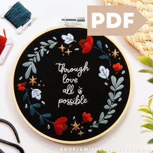 Op de afbeelding: Een zwarte borduurring met een bloemenkransontwerp en de tekst "through love all is possible". De ring is omgeven door borduurgaren in rood, blauw en groen.