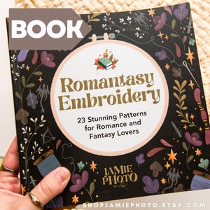 May include: A square book titled Romantasy Embroidery with a black cover featuring colorful illustrations of flowers, stars, and swords. The center shows a circular embroidery hoop design with the text 23 Stunning Patterns for Romance and Fantasy Lovers.