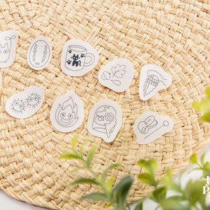 May include: A set of 10 small, white, sticker-like decals with black line drawings of various characters and objects. The decals are arranged on a woven, straw-like surface.
