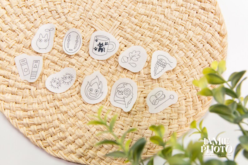 May include: A set of 10 small, white, sticker-like decals with black line drawings of various characters and objects. The decals are arranged on a woven, straw-like surface.