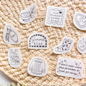 May include: A collection of ten white stickers with black line art illustrations including a motel keychain with 1821, a stamp reading we didn't even kiss, a heart with S+K, socks, candy hearts with ILYA and SHANE, and a jersey with ROZANOV 81.