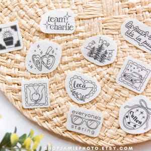 May include: A collection of nine white fabric patches featuring black line art designs including a cactus, a heart locket with BE, a wolf inside a heart with loca, a car, hands holding an apple, glasses with everyone's staring, and text.