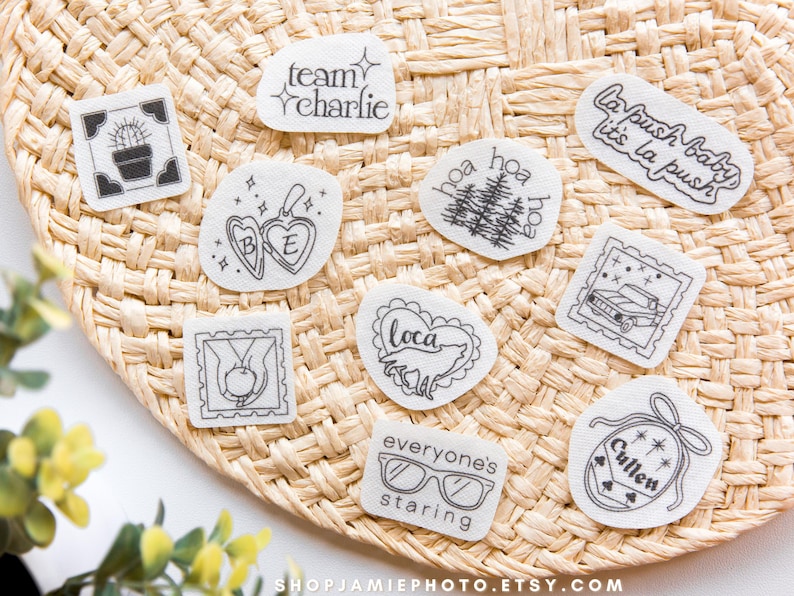 May include: A collection of nine white fabric patches featuring black line art designs including a cactus, a heart locket with BE, a wolf inside a heart with loca, a car, hands holding an apple, glasses with everyone's staring, and text.