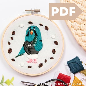 May include: Embroidered hoop art featuring a teal blue bird wearing a bow tie and glasses, surrounded by coffee beans. The hoop is made of wood and has a white background.
