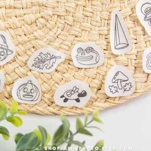 May include: A collection of ten white stickers featuring black line art illustrations of a jack-o'-lantern, a frog, a turtle, a mushroom, a lantern, a leaf, a cone, and other whimsical characters arranged on a woven straw surface.