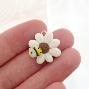 May include: A daisy charm with a white flower, brown center, and a yellow and black bee. The charm has a silver loop for attaching to jewelry. The charm is likely made of resin or a similar material.