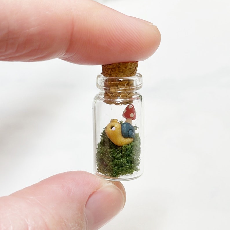 May include: A miniature glass bottle with a cork stopper, containing a tiny snail figurine on a bed of green moss, with a small red and white mushroom. The snail has a yellow body and a blue shell.