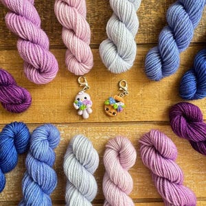 May include: An overhead shot of colorful yarn skeins in shades of purple, pink, blue, and gray. Two small charm pendants are also visible. The yarn is arranged on a wooden surface, creating a visually appealing display for crafting projects.