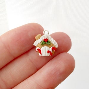 May include: A miniature gingerbread house charm with a silver loop for hanging. The house features a brown base, white frosting, red and green holly, and candy cane accents. The door is white with a red doorknob.
