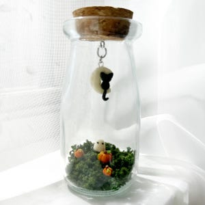 May include: A clear glass bottle with a cork stopper, featuring a Halloween-themed terrarium. Inside, there's a miniature landscape with green moss, pumpkins, and a small ghost. A black cat charm hangs from the cork.