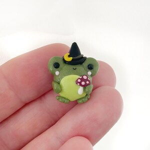 May include: A handcrafted green frog figurine wearing a black witch hat with a yellow crescent moon. The frog holds a mushroom with a red cap and white spots. The figurine is made of clay.