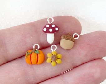 Könnte beinhalten: Eine Sammlung von Miniatur-Anhängern mit herbstlichen Designs. Enthält einen roten Pilz mit weißen Punkten, einen orangefarbenen Kürbis, eine gelbe Sonnenblume und eine beige Eichel. Jeder Anhänger hat eine silberne Schlaufe zum Befestigen.