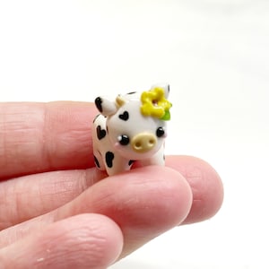 Puede incluir: Una figura de vaca blanca en miniatura con manchas negras y una flor amarilla en la cabeza. La vaca tiene una nariz y mejillas rosadas, y marcas negras en forma de corazón. La figura se sostiene en una mano.
