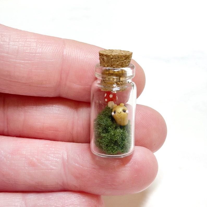 May include: A miniature glass bottle with a cork stopper, containing a small scene. Inside, there's green moss, a red mushroom with white spots, and a tiny, tan-colored creature with black eyes. The bottle is held in a person's hand.