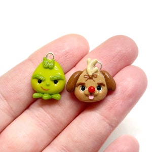 May include: Two handmade polymer clay charms. One is a green Grinch-like character with a bow and a mischievous expression. The other is a brown dog with a red nose, antlers, and a lolling tongue. Both charms have silver-colored loops.