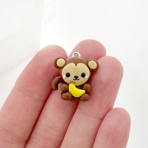 May include: A brown monkey charm with a light brown face and ears, holding a yellow banana. The charm has a silver loop at the top for attaching to a necklace or bracelet. The monkey has black eyes and a small black nose.