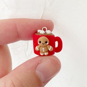 May include: A miniature red mug charm with a gingerbread man and marshmallows. The mug has a handle and is filled with brown liquid, topped with white marshmallows. The gingerbread man has white icing details and is smiling.