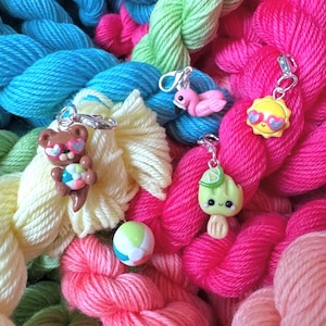 May include: A collection of colorful yarn skeins with various summer-themed charms. The charms include a bear with sunglasses, a flamingo, a sun with heart-shaped sunglasses, and a lime popsicle. A small beach ball is also present.