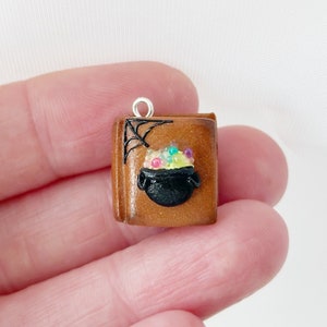 May include: A miniature brown book charm with a black cauldron filled with colorful beads. A black spiderweb design is on the upper left corner. The charm has a silver loop for attaching to a necklace or bracelet.