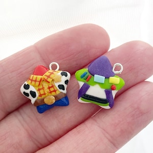 Könnte beinhalten: Zwei sternförmige Charms mit Figuren aus dem Animationsfilm Toy Story. Ein Charm zeigt Woody in einem gelben Hemd, blauen Hosen und einem braunen Gürtel. Der andere Charm ist Buzz Lightyear, mit einem lila Helm und einem grün-weißen Anzug.