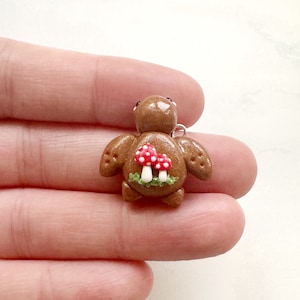 May include: A brown turtle charm with a mushroom design on its shell. The mushrooms have red caps with white spots and are set on a bed of green and white grass. The charm has a silver-colored loop for attaching to a necklace or bracelet.