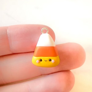 May include: A colorful candy corn charm with a smiling face and pink cheeks. The charm features a white top, orange middle, and yellow bottom, with a silver loop for attaching to jewelry or other crafts.
