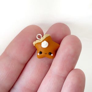 Op de afbeelding: Een handgemaakte stervormige bedel van oranje polymeerklei met een lachend gezicht. De bedel heeft een bruin topje en een toefje witte crème. Het heeft een zilverkleurige lus om aan te bevestigen. De bedel wordt in een hand gehouden.