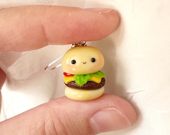 May include: A miniature, smiling hamburger charm with a bun, patty, lettuce, tomato, and cheese. The charm is held by a silver-colored earring hook. The burger has a cute, cartoonish design.