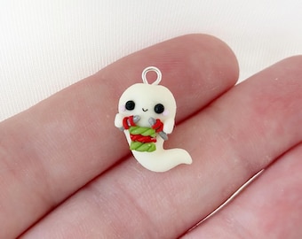 May include: A small, off-white ghost charm with a silver loop for hanging. The ghost has black eyes, a small smile, and is wearing a red and green scarf. The charm is held in a person's hand.