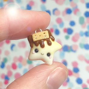 May include: A star-shaped charm with a smiling face, made of off-white clay, topped with brown chocolate and a small biscuit. The charm has black eyes and pink cheeks. A silver loop is attached for hanging. The text "TheLittlestCharmCo" is visible.