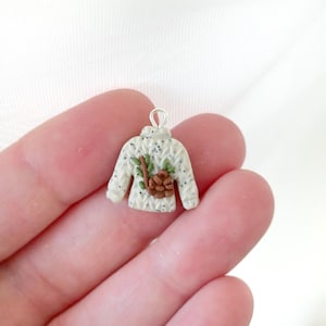 Könnte beinhalten: Ein kleiner, cremefarbener Pullover-Anhänger mit einer silbernen Schlaufe. Der Pullover ist mit einem braunen Tannenzapfen und grünen Blättern verziert. Der Pullover hat schwarze Sprenkel und ein strukturiertes Strickmuster.
