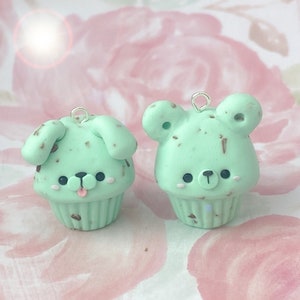 Puede incluir: Dos colgantes de cupcake verde menta, uno con forma de perro y otro de oso. Ambos tienen motas marrones y lazos plateados para colgar. El colgante de perro tiene orejas caídas y una lengua rosa.