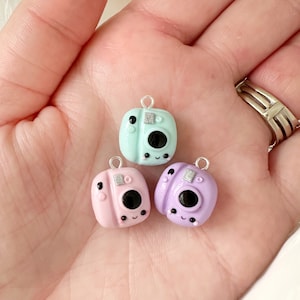 May include: Three miniature camera charms in pastel shades of light blue, pink, and lilac. Each has a black lens, silver detailing, and a smiling face. The charms are small, with a loop for attaching to jewellery or crafts.