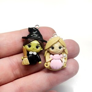 May include: Two miniature polymer clay figurines: a witch with a black hat and broom, and a princess in a pink dress. Both have smiling faces and are designed with fine details. Each figure has a silver-colored loop at the top for hanging.