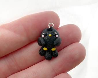 May include: A small, handmade black bird charm with a silver-coloured loop for hanging. The bird has a yellow beak and feet, and pink cheeks. The charm is approximately 2.5 cm in height and is made of a smooth, possibly polymer clay material.
