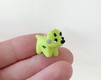 May include: A small, lime green dog charm with black accents and a silver-coloured loop. The charm has a stitched design on its side and a grey circle on its cheek. The dog's nose and eyes are black.