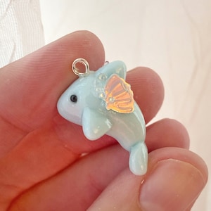 May include: A light blue dolphin charm with a silver loop for hanging. The dolphin has a small, iridescent, orange-yellow fin and small, clear, iridescent bubbles. The dolphin's eye is black.