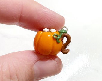 Könnte beinhalten: Ein miniaturförmiger, orangefarbener Kürbis-Tassen-Anhänger mit braunem Griff und grünen Rankendetails. Die Tasse ist mit heißer Schokolade und zwei weißen Marshmallows gefüllt. Eine silberne Schlaufe ist zum Aufhängen angebracht.