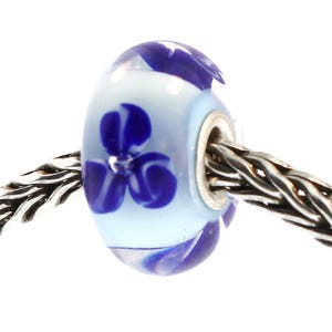 May include: A glass bead with a light blue base and a dark blue floral design. The bead is cylindrical with a silver-colored metal insert. The bead is strung on a silver-colored chain.