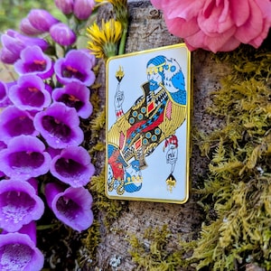 May include: A rectangular enamel pin featuring a king from a deck of cards, with a gold border. The king is depicted with a blue beard and crown, holding a torch. The pin is set against a backdrop of flowers and moss.