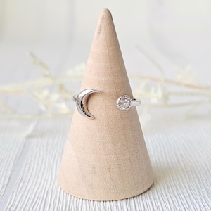 Könnte beinhalten: Ein silberner Ring mit einem Halbmond-Design und einem runden, klaren Edelstein. Der Ring wird auf einem hellbraunen, kegelförmigen Holzständer vor einem weichen, verschwommenen Hintergrund präsentiert.