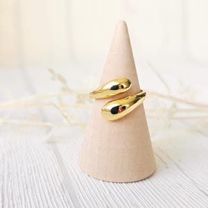 Könnte beinhalten: Ein goldfarbener Ring mit Wickeldesign. Der Ring hat eine glatte, polierte Oberfläche und wird auf einem hellrosa kegelförmigen Ständer präsentiert. Der Hintergrund zeigt eine weiße Holzoberfläche und weiche, verschwommene Details.