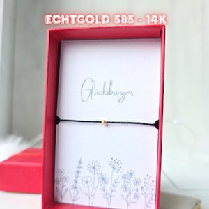Könnte beinhalten: Ein Armband mit einer goldenen Perle an einem schwarzen Band wird in einer roten Schachtel präsentiert. In der Schachtel befindet sich eine weiße Karte mit dem Wort "Glücksbringer" und floralen Illustrationen. Der Text "ECHTGOLD 585 - 14K" steht oben.