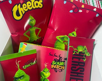 Może przedstawiać: Pudełko prezentowe wypełnione przekąskami z motywem Grincha. Pudełko zawiera torebki Cheetos, Sour Patch Kids, mieszankę gorącej czekolady Swiss Miss i batonik czekoladowy Hershey's. Opakowanie jest czerwono-zielone ze świątecznymi lampkami i grafiką Grincha.