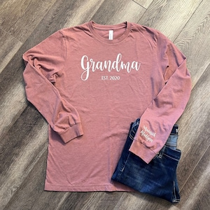 Pu&ograve; includere: Una maglietta a maniche lunghe color rosa polvere con la scritta "Grandma Est. 2020" stampata in bianco sul davanti. La manica sinistra ha la scritta "Maverick Madison" stampata in bianco.