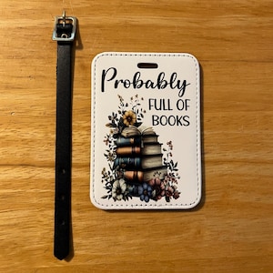 May include: A white luggage tag with the text "Probably FULL OF BOOKS" and a stack of books with floral accents. A black leather strap with a silver buckle is attached to the tag.