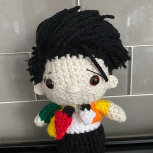 May include: A crocheted doll with black hair, a white shirt, a colorful jacket, and black pants. The doll has brown eyes and a black bow tie.
