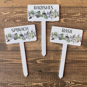 May include: Three white garden markers with text and illustrations. The markers read "RADISHES", "SPINACH", and "BASIL". Each marker has a rectangular top with a white stem. The illustrations feature gardening tools and plants.