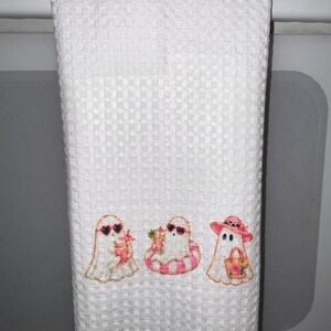 May include: A white waffle-weave kitchen towel featuring an embroidered design of three ghosts. The first ghost wears heart-shaped sunglasses and holds a drink. The second ghost sits in a pink striped inner tube with a drink. The third ghost wears a pink sun hat and carries a tote bag.