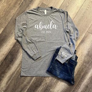 Pu&ograve; includere: Maglietta grigia a maniche lunghe con il testo "abuela est 2024" in bianco. La maglietta &egrave; abbinata a un paio di jeans blu.